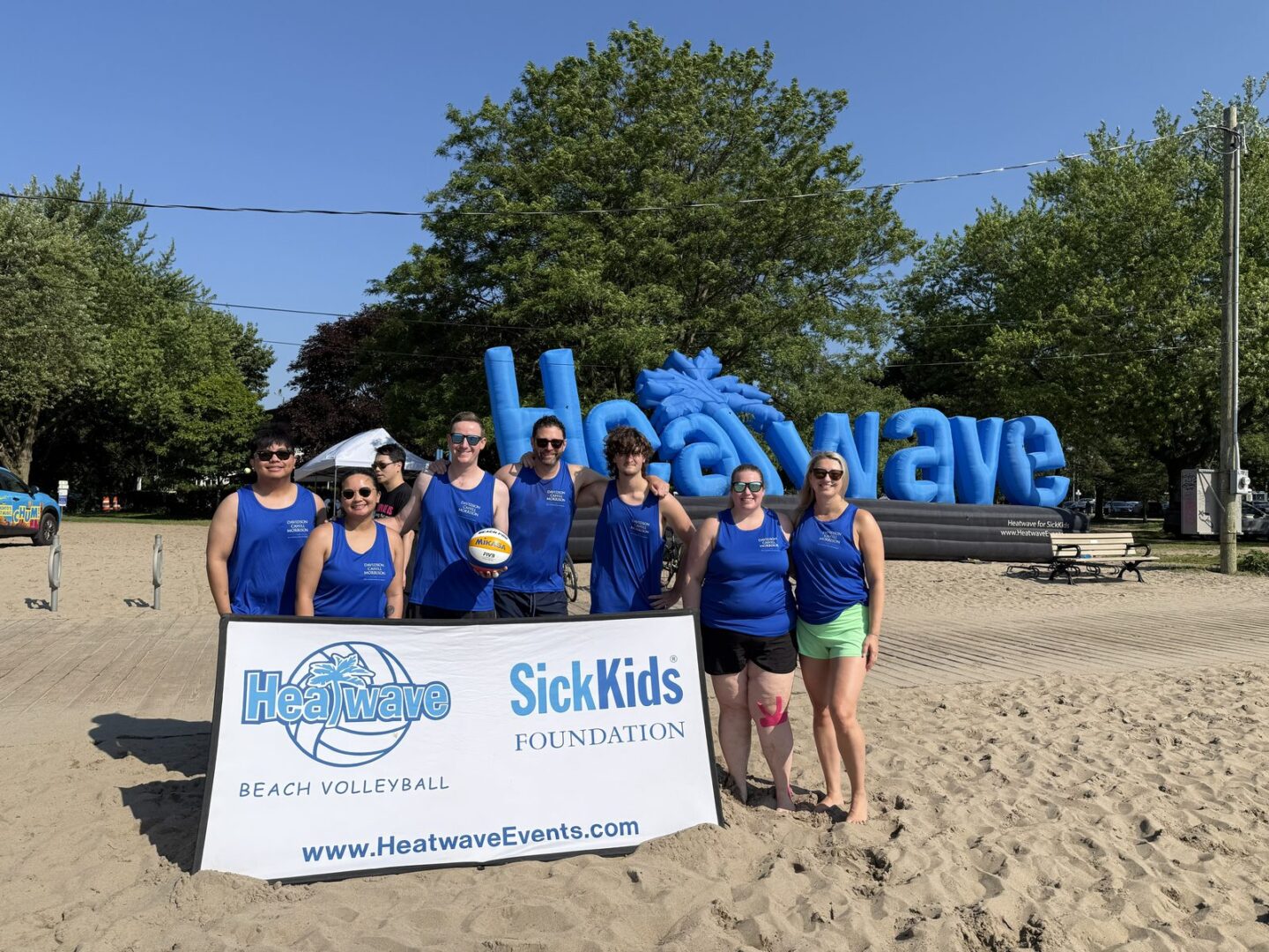 Playing for SickKids at the 2026 Heatwave Beach Volleyball Tournament | Davidson Cahill Morrison LLP The Davidson Cahill Morrison LLP team in matching blue jerseys on the sand at Woodbine Beach, holding a volleyball in front of the Heatwave for SickKids banner.
